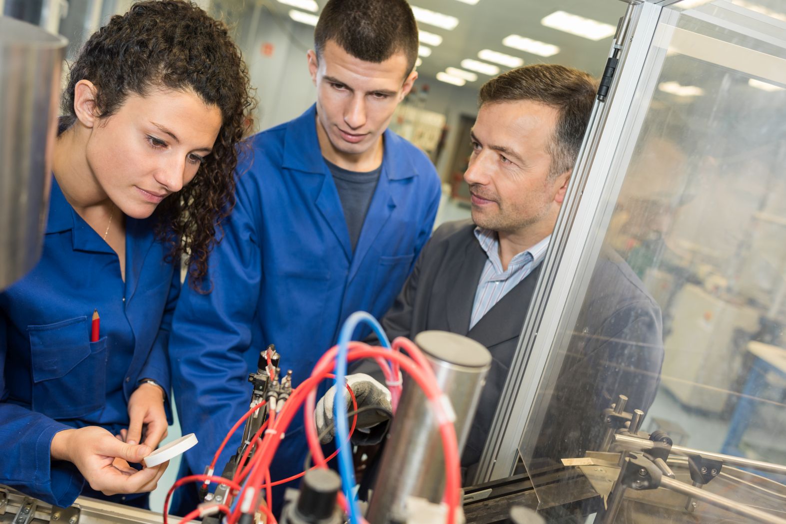 Drei Personen stehen in einer Werkhalle vor einer Maschine. Im Vordergrund sind Kabel und Maschinenteile sichtbar.