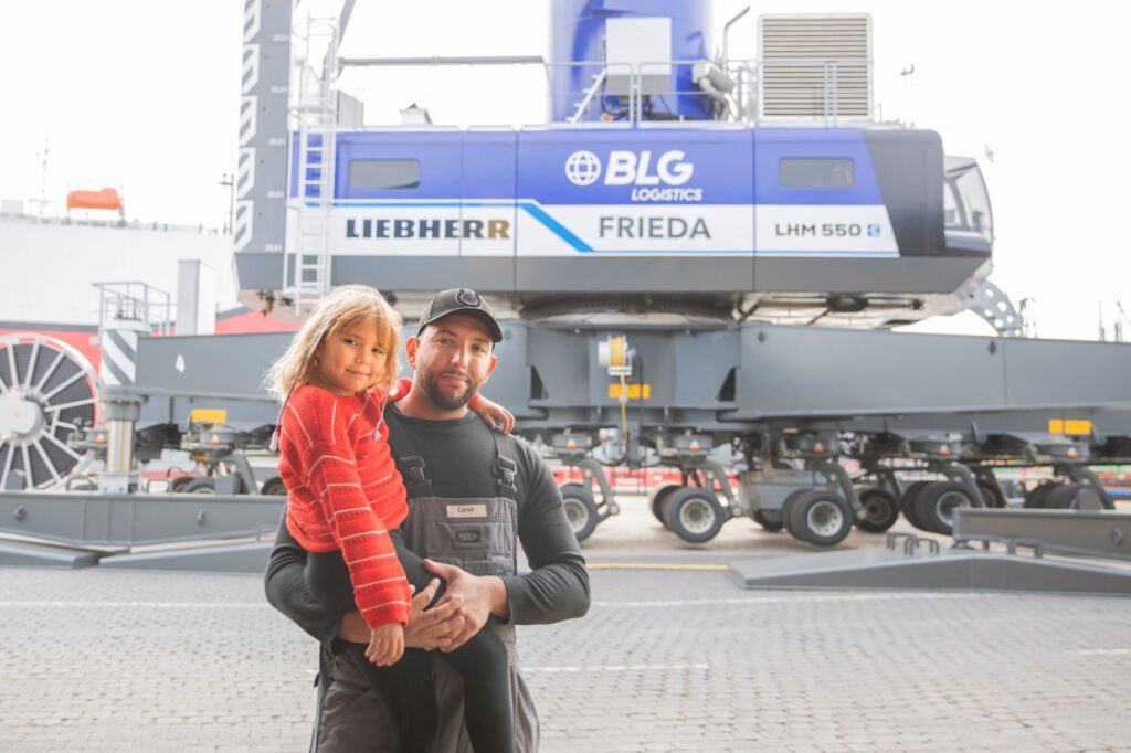 Ein Mann hält seine Tochter im Arm. Beide blicken direkt in die Kamera. Im Hintergrund ist ein großer Hafenkran zu sehen.