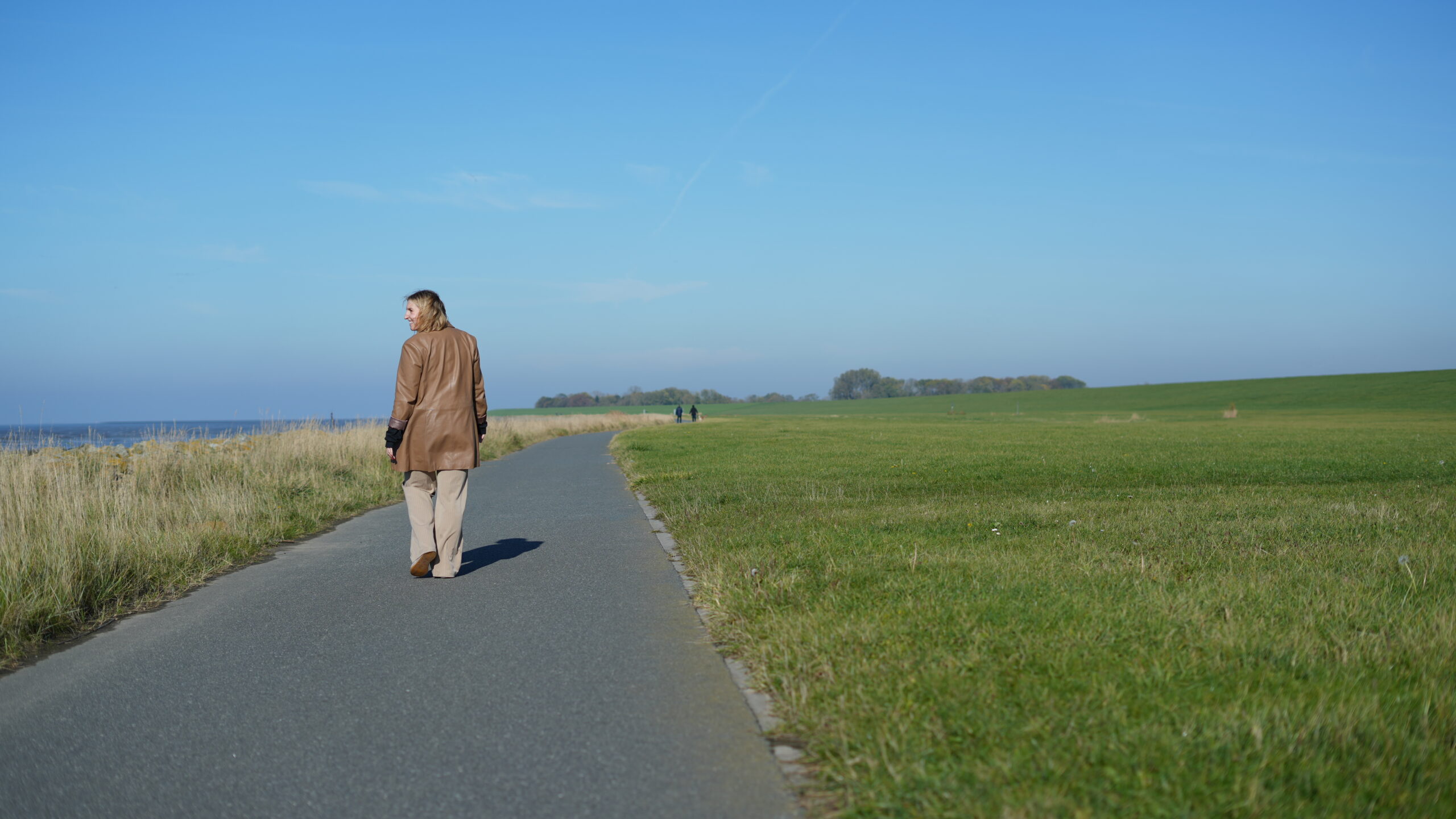 Person geht einen Weg zwischen Wasser und Deich entlang.