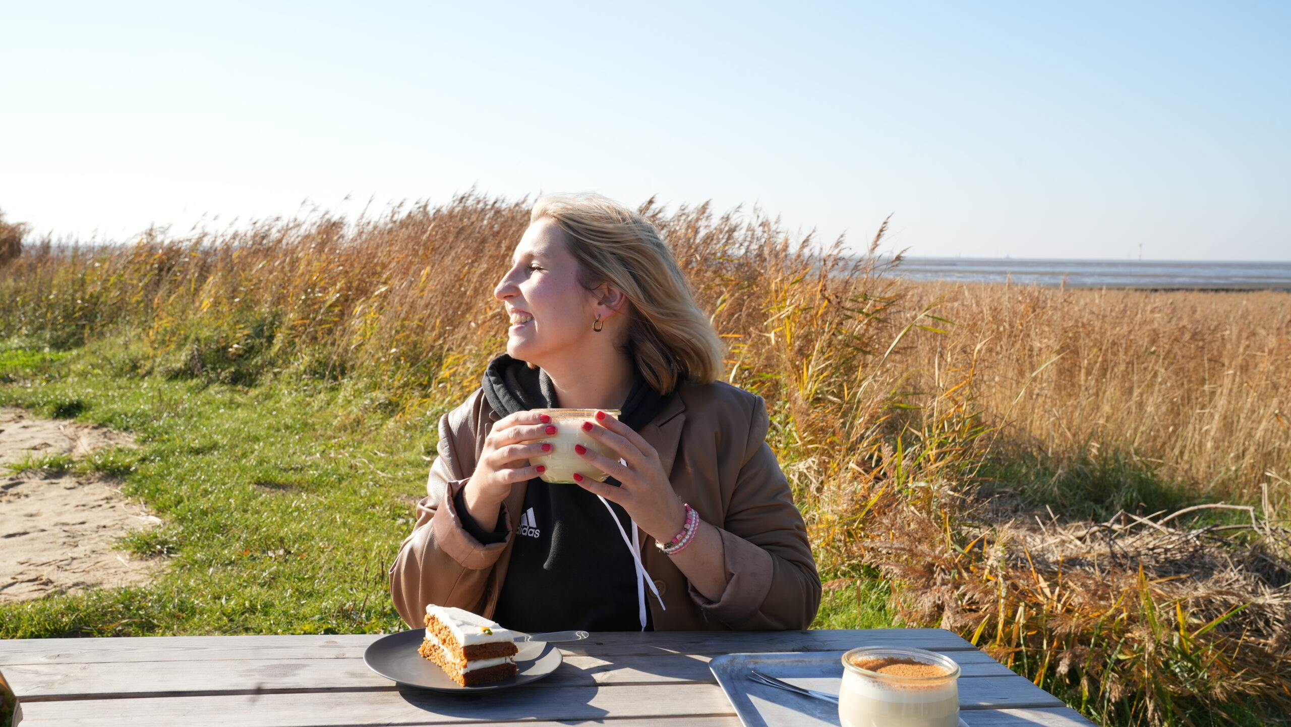 Person sitzt an einem Tisch am Meer mit Getränk und Kuchen.