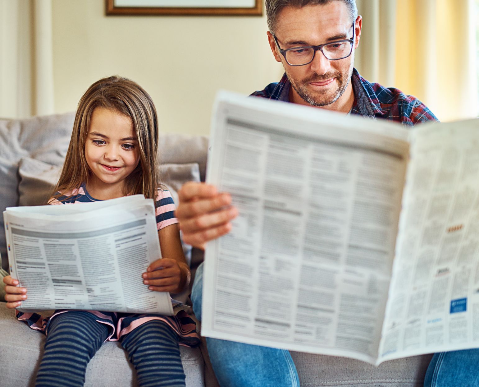 Ein Vater und seine Tochter lesen Zeitung, während sie nebeneinander auf dem Sofa sitzen.