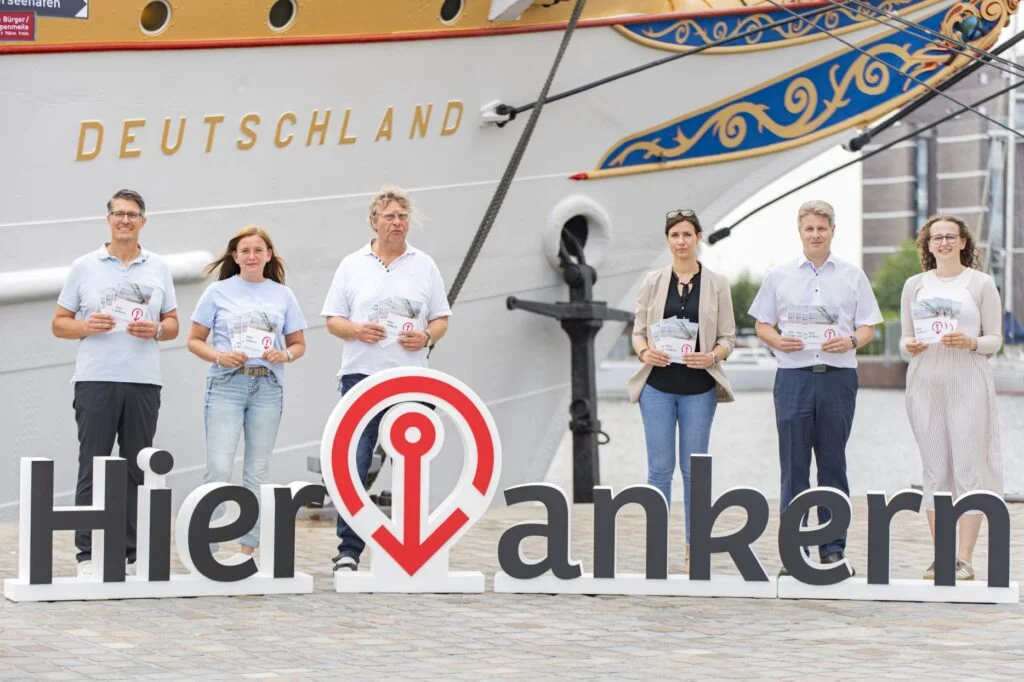 Vor der „Schulschiff Deutschland“ in Bremerhaven präsentieren Jan Rathjen (Nordsee-Zeitung), Jenny Bergmann (im-jaich), Wolfgang Turkowsky (Hotel an der Karlstadt), Fenja Eichstädt (SNW Hotels), Piet Rothe (Hotels Adena und Amaris) und Rieke Nagel (Atlantic Hotel Sail City) die neuen „Hier ankern“-Flyer, mit denen Bremerhavener Hotels die Fachkräftekampagne künftig aktiv unterstützen.