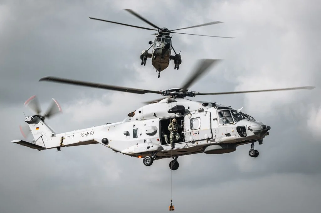 Ein NH90 „Sea Lion“ (Vordergrund) der Nordholzer Marineflieger im Flug. Neue Wartungshallen für die Instandhaltung sind im Industriegebiet Wanhöden neben dem Stützpunkt geplant; General Atomics Europe hat sich dort eine Fläche gesichert.